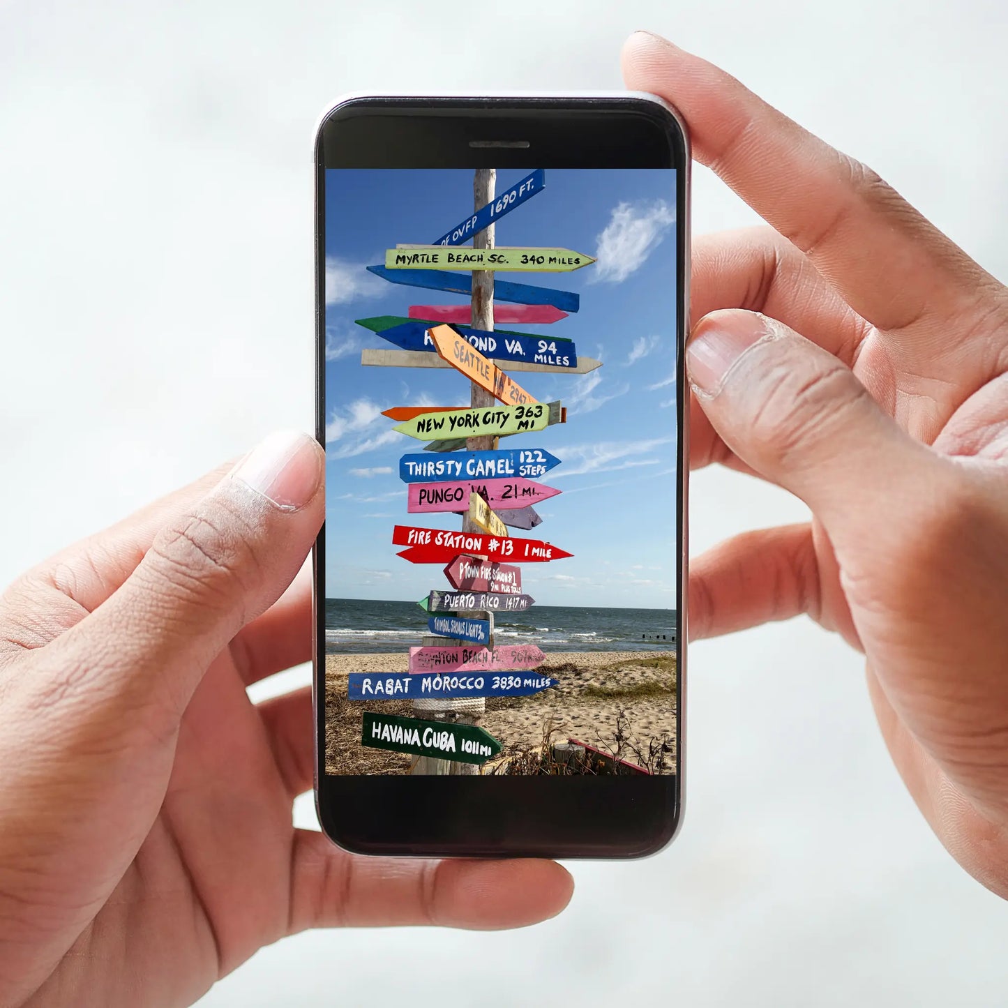 Person holding a smartphone displaying colorful directional signs on a beach, indicating various destinations.